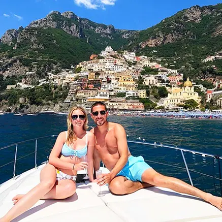 A couple relaxes on the sun deck of a private boat charter with the colorful village of Positano and the steep coastline as the background during a full day Amalfi Coast tour