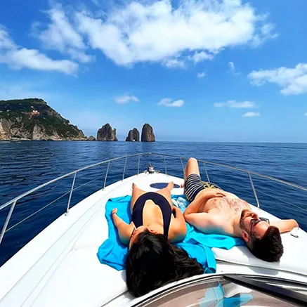 A couple sunbathing on the bow of a private motor yacht during a full day Capri boat charter with the Faraglioni rocks in the background.